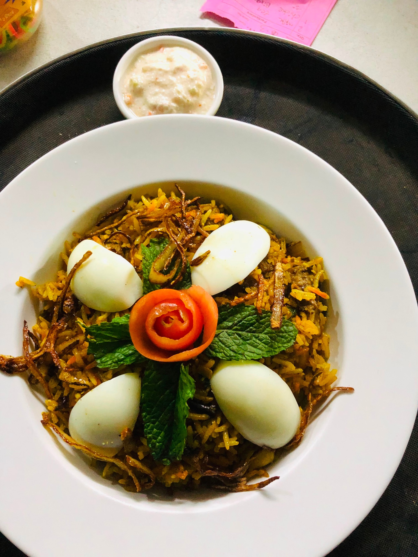 Bowl of egg biryani topped with boiled eggs, mint, fried onions and a tomato rose, with raita